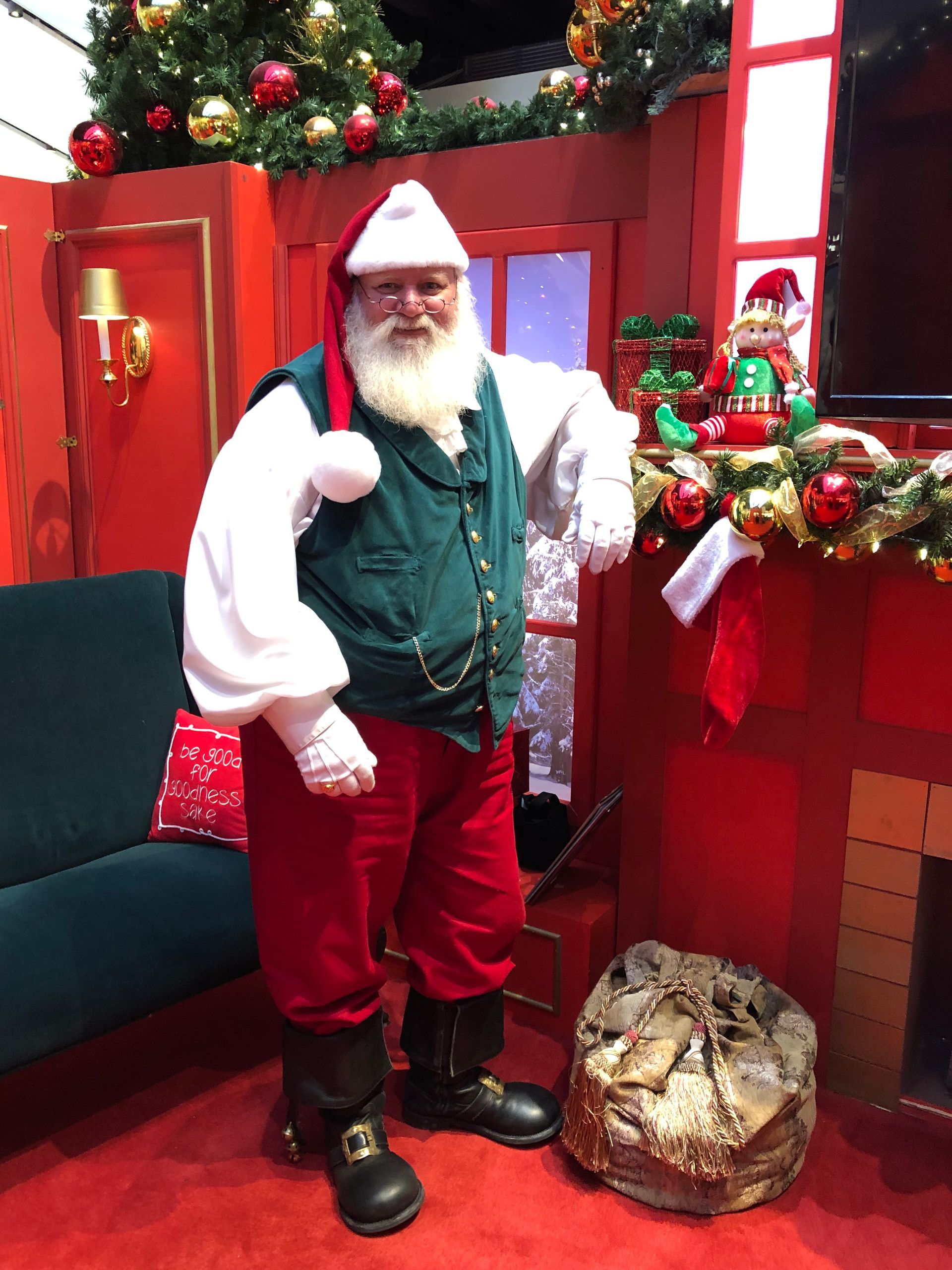 Santa Claus in a red room, leaning on a fireplace. He wears a green vest, red pants, and a white beard.