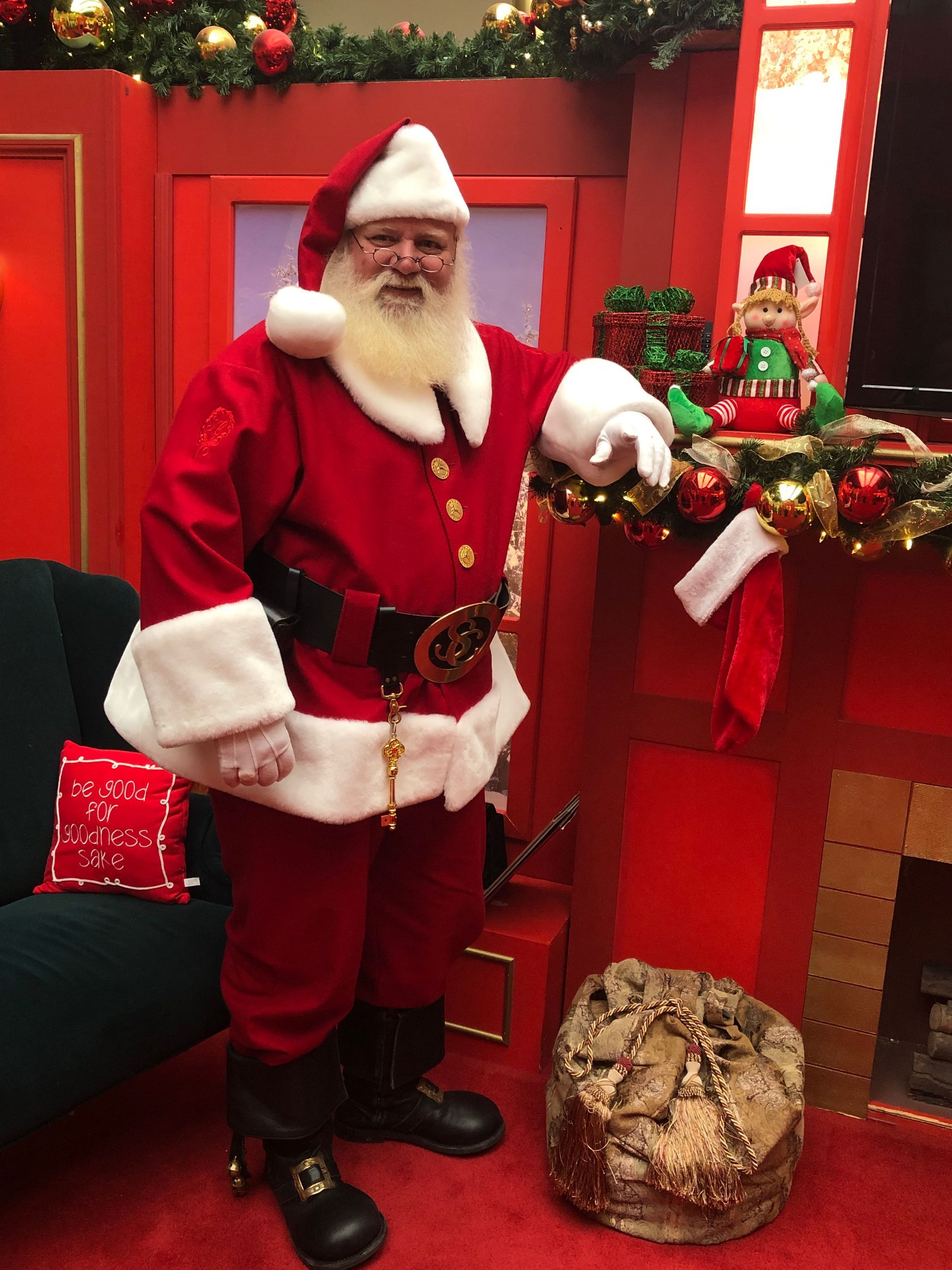 Santa Claus in red suit, white beard, smiling. Standing by a fireplace in a red room, decorated for Christmas.