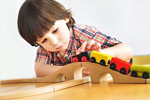 Child leaning over a wooden toy train track, watching a red and yellow toy train