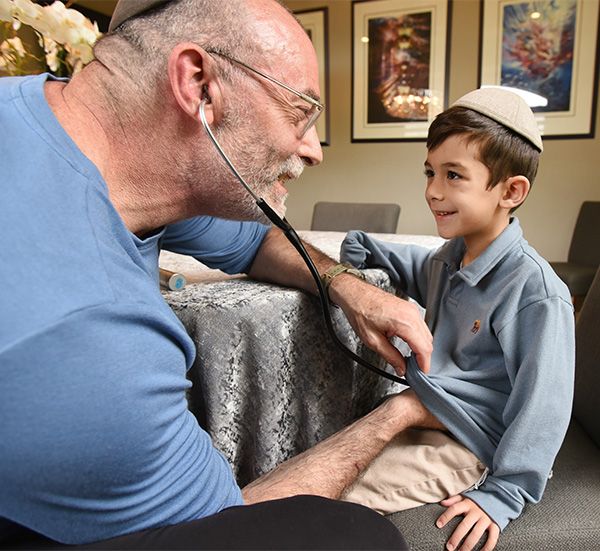 Doctor listens to a boy's chest with a stethoscope. Both are wearing yarmulkes; the setting appears to be a home.