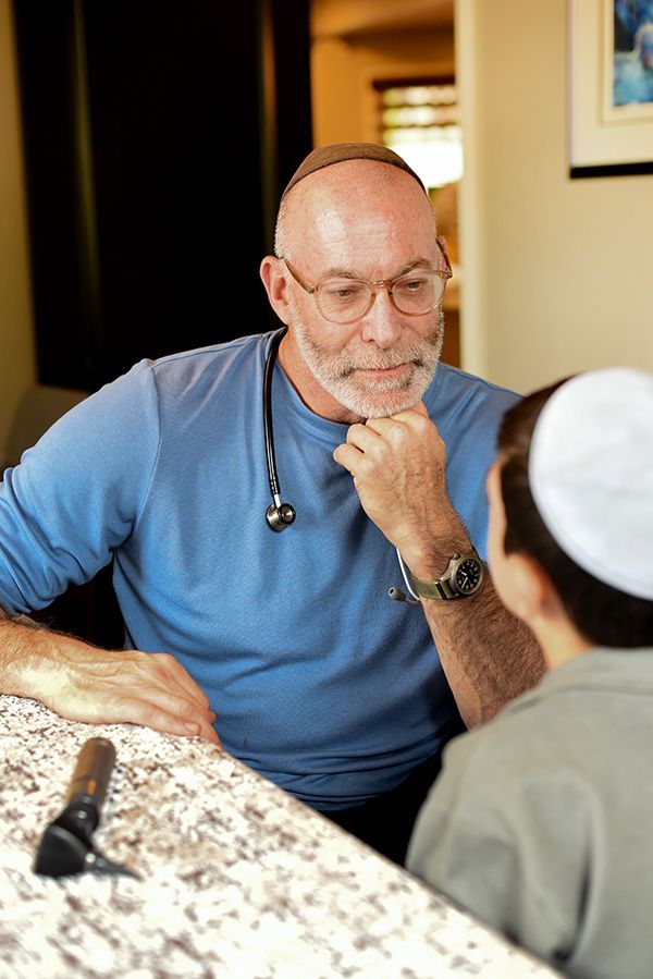 Doctor in blue shirt, stethoscope, and yarmulke consults with a boy wearing a yarmulke at a table; otoscope visible.