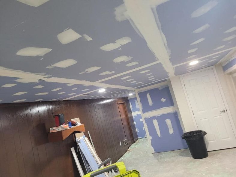 Basement renovation in progress; drywall on ceiling and walls, blue and white, with a door and trash can.