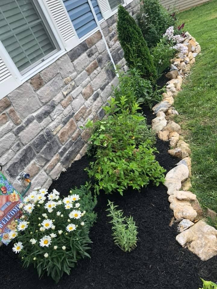 Stone house with a flower bed of plants and daisies edged with rocks and black mulch.