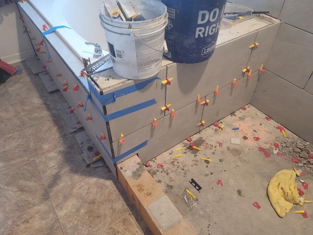 Tiling in progress: large gray tiles installed with spacers on a bathroom ledge. Tools and buckets present.