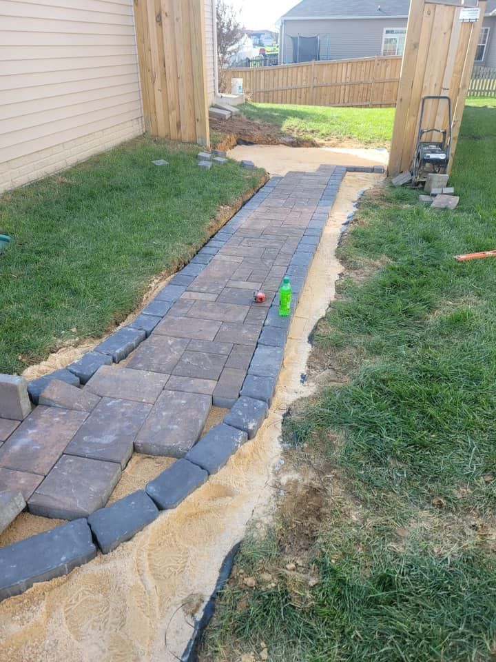 A partially completed brick walkway, between a house and fence, with sand base and landscaping edging.