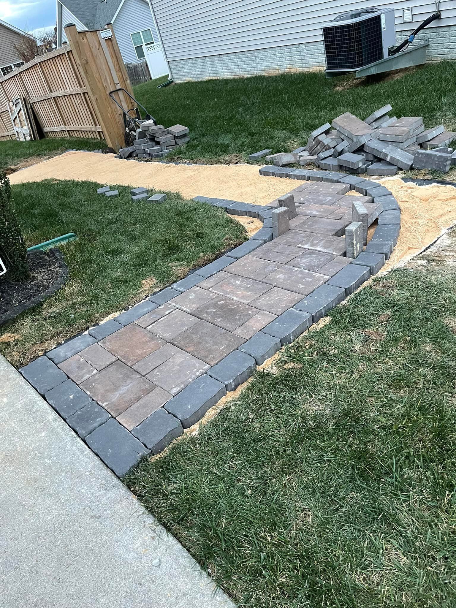 A partially completed brick walkway with a dark border, surrounded by grass and sand, in front of a house.