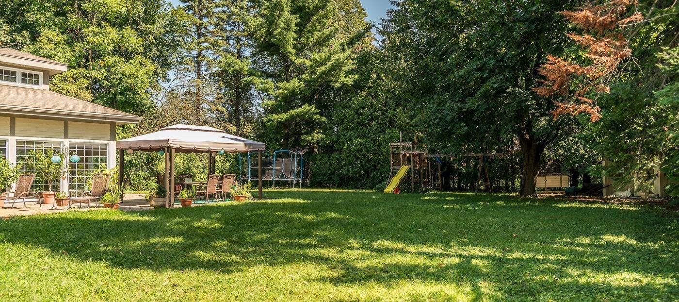 Lush backyard with gazebo, swing set, and green grass. Trees frame the sunny landscape near a house.