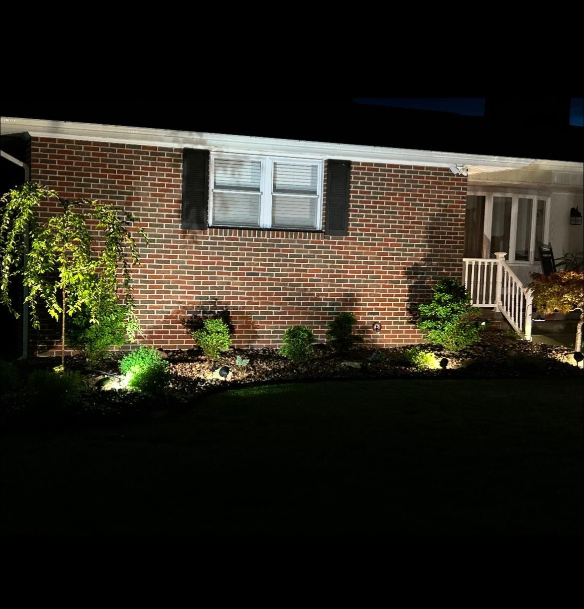A brick house with illuminated landscaping at night, including trees, bushes, and a pathway to the front door.