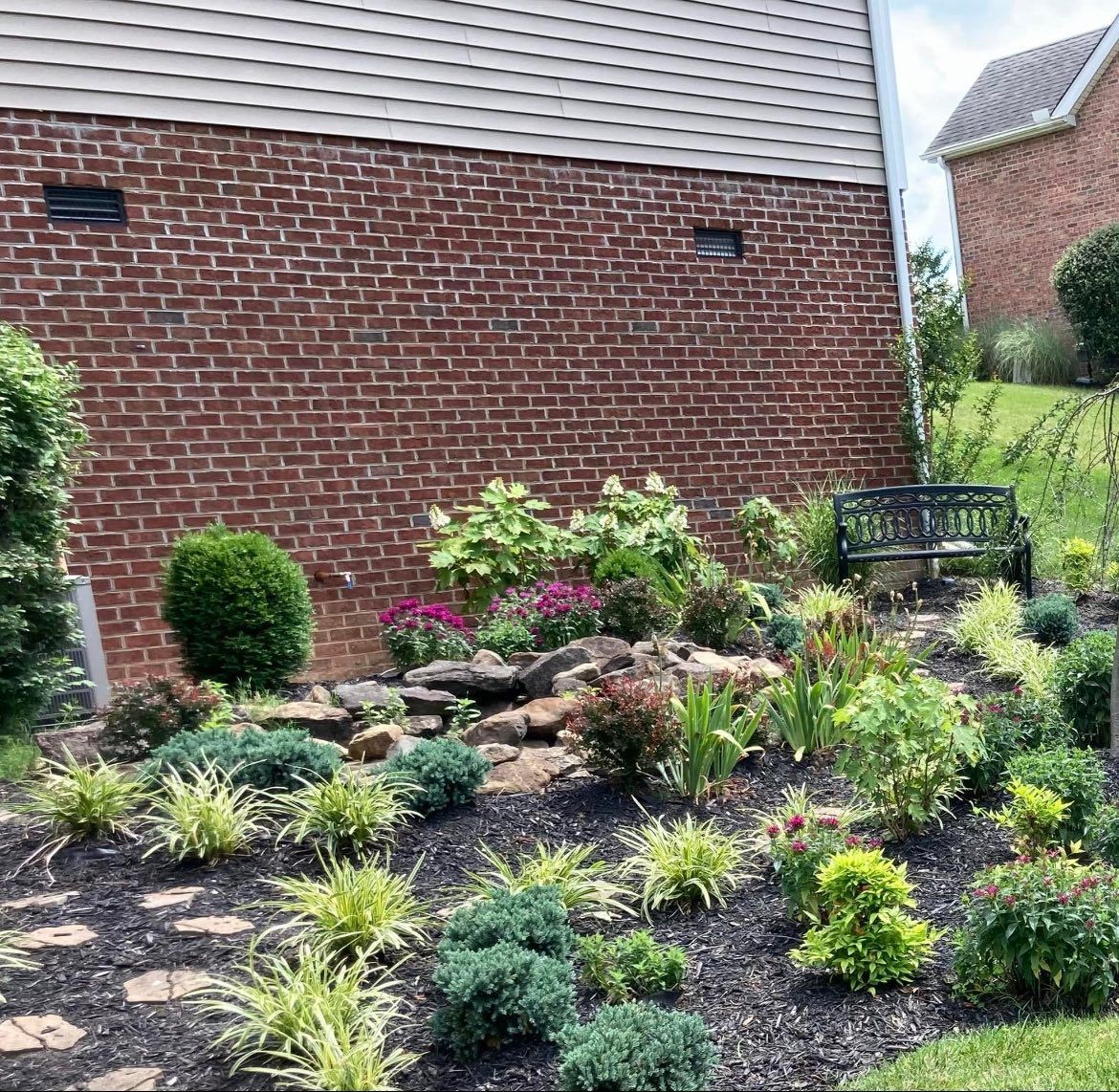 Garden bed with various plants and a bench against a brick wall.