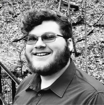 Man with glasses and a beard smiling, wearing a button-down shirt. Outdoors with foliage background.