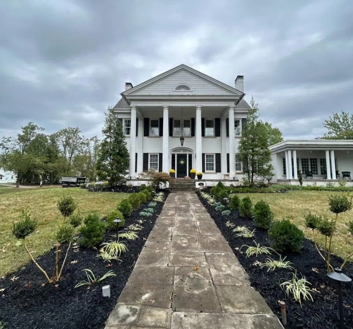 White colonial home with large pillars, a path, and manicured landscaping under a cloudy sky.