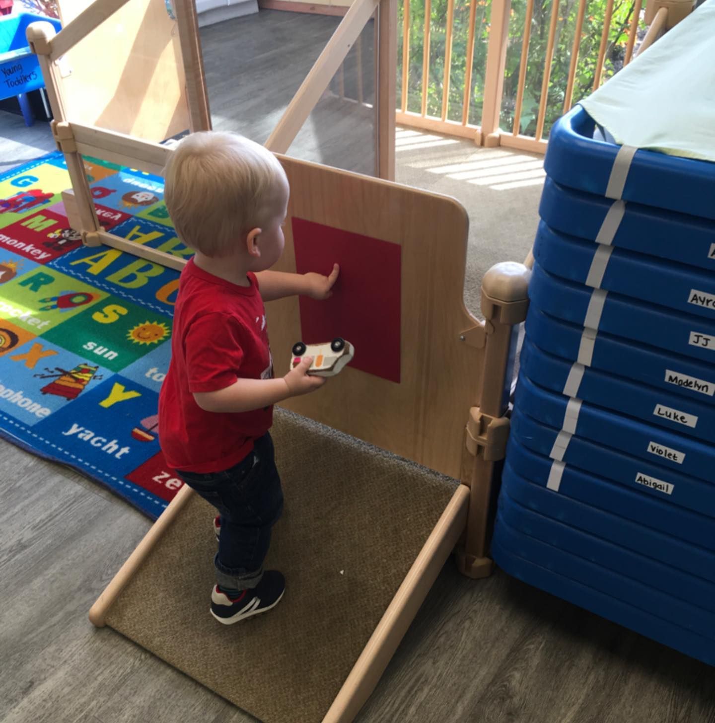 A toddler in red shirt plays with a toy car near a red square on a wooden play structure.