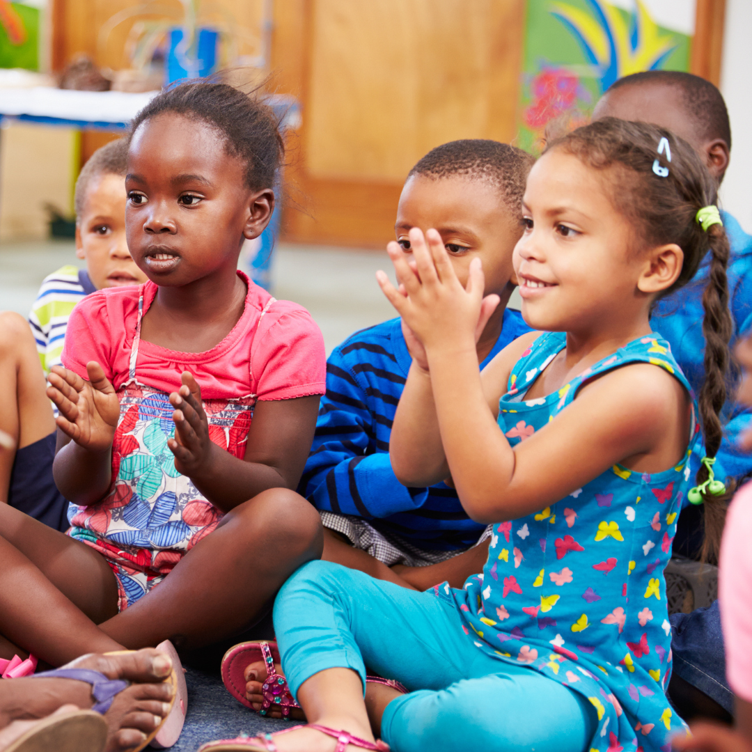 Smiling baby wearing a brown knit hat with two pom-poms, indoors.
