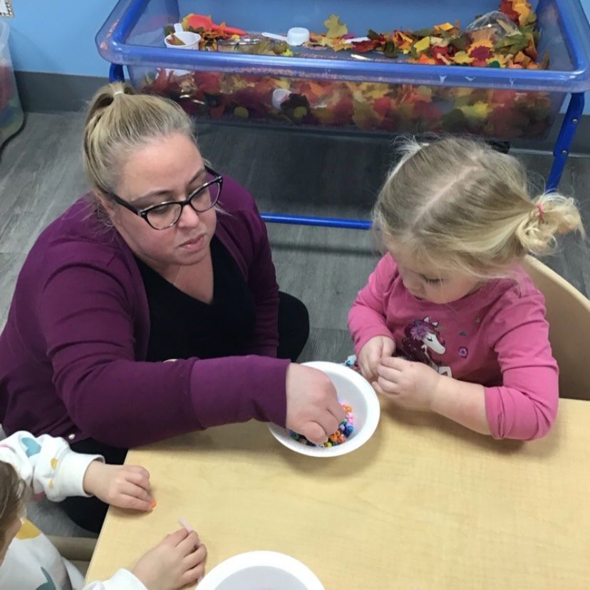 Woman and child at a table, sorting items from a bowl; fall-themed sensory bin in background.