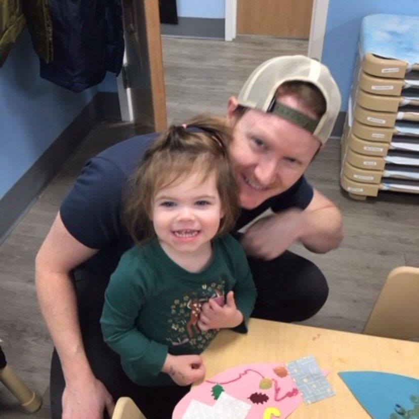 Man and child smiling at the camera, seated at a small table decorated with craft supplies in a classroom.