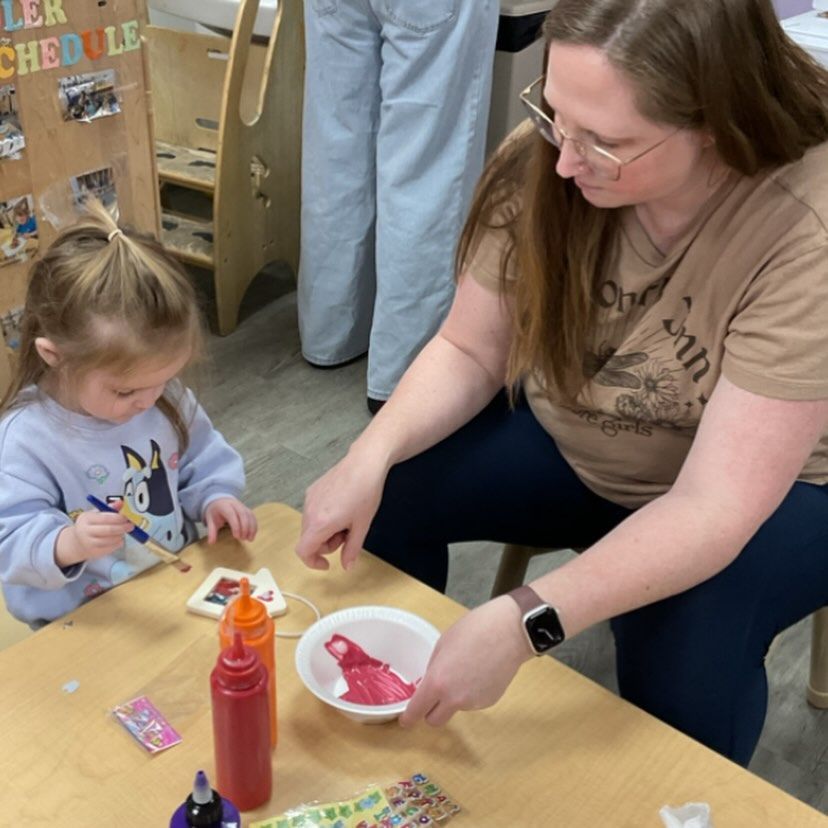 Child and adult painting at a table in a classroom. The child has a paintbrush and is focused.