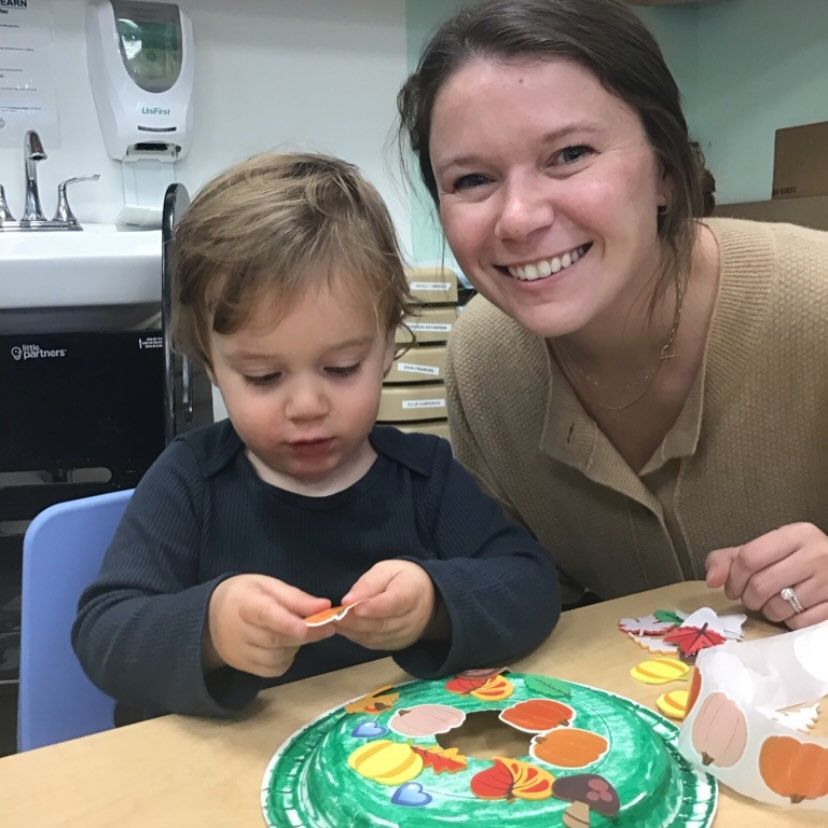Woman and young child making a fall-themed craft. The child examines a sticker, while the woman smiles.