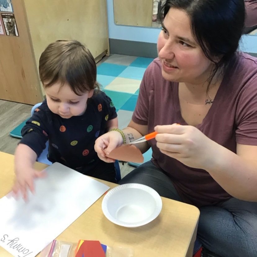 A person helps a young child with a craft at a table; child touches paper.