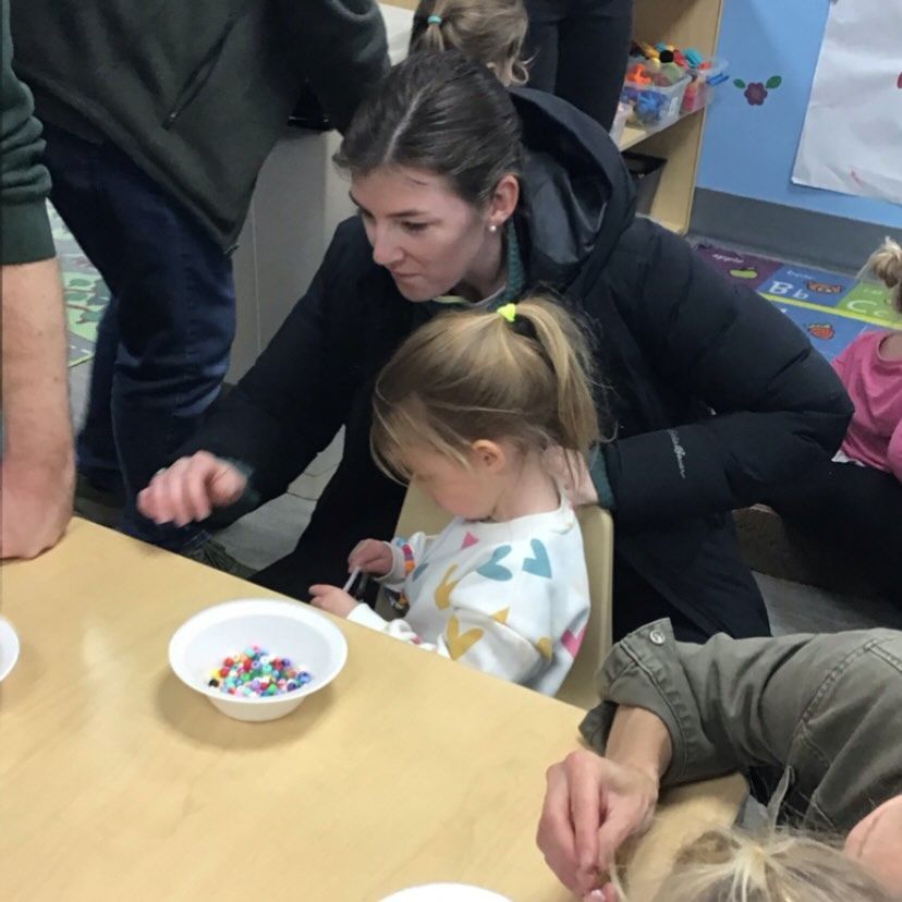 Woman assisting a toddler with beads at a table in a classroom.