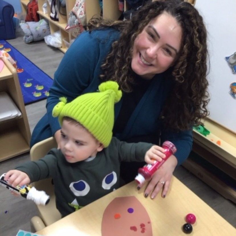 Woman with curly hair smiles next to child in green hat, painting at a table.