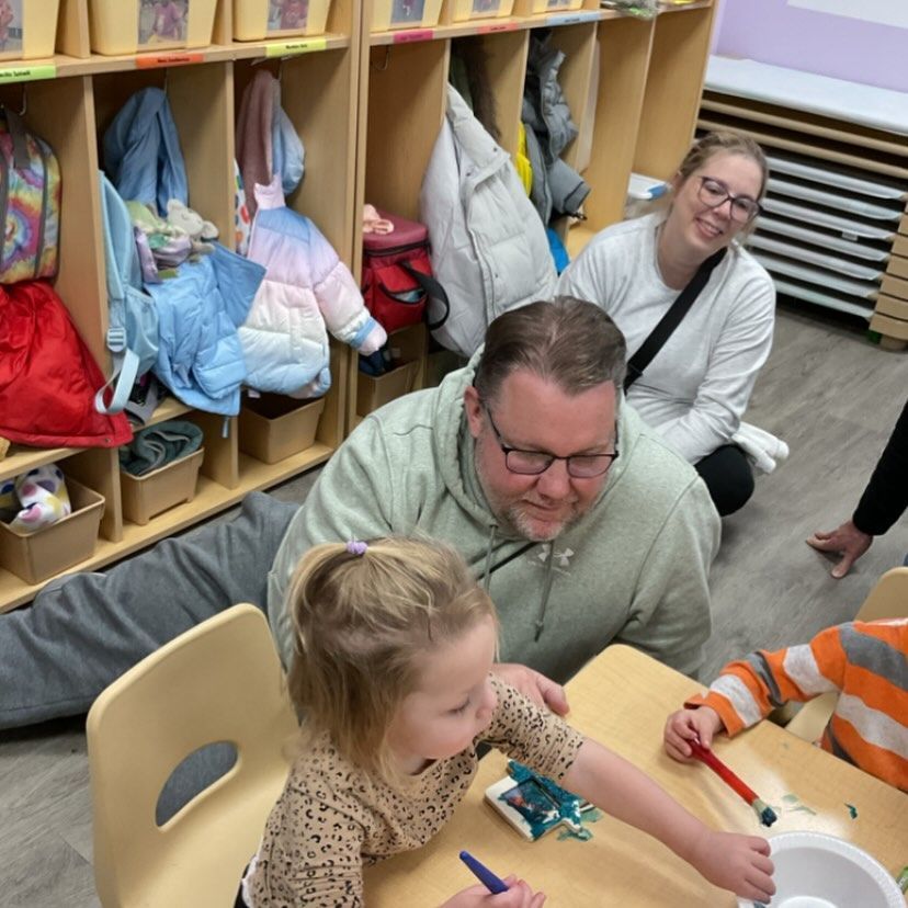 Children and adults at a table painting in a classroom. A woman and man observe.