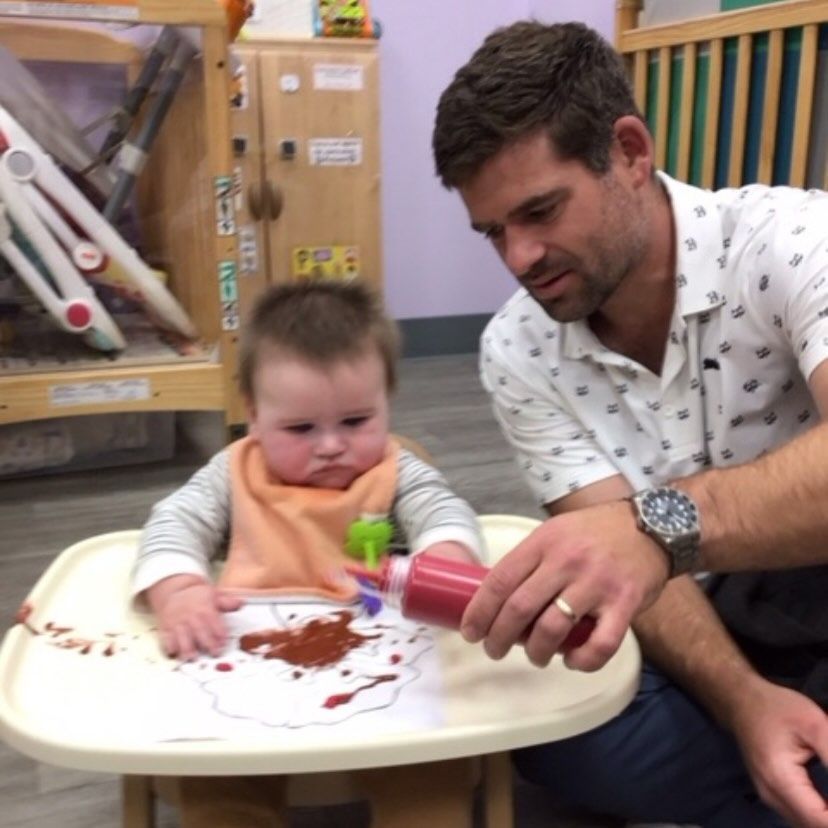 A baby in a highchair splatters red paint while a man watches, both in a playroom setting.