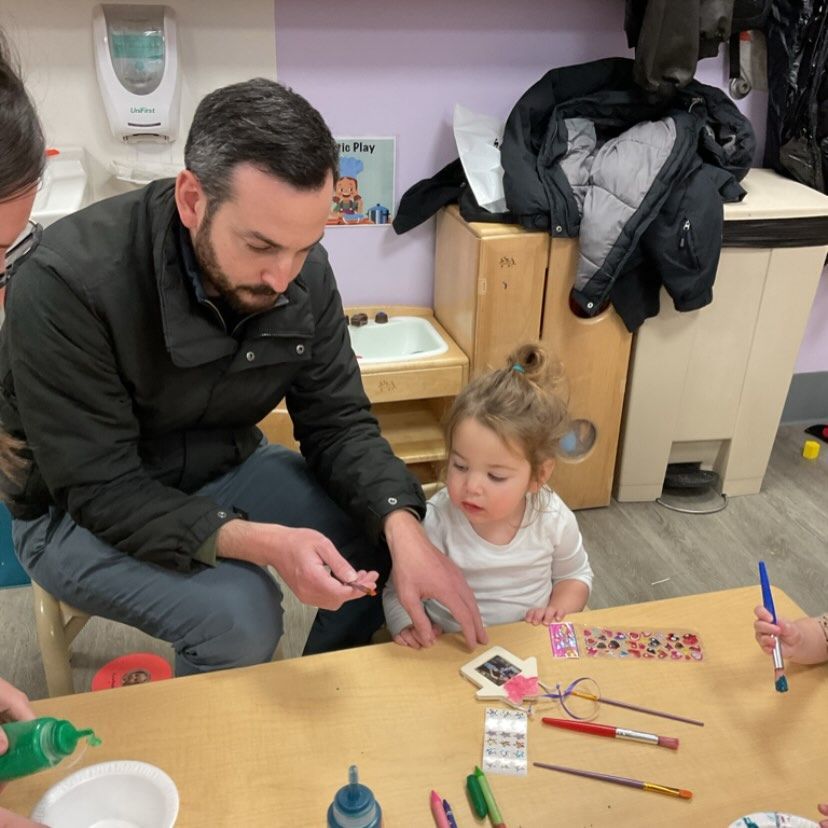 Man and toddler at a table crafting. The man points at an item. Child looks on attentively.