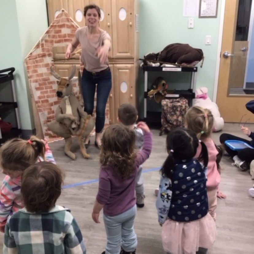 Woman leading children in activity; they are in a room with a decorative brick wall and a stuffed deer.