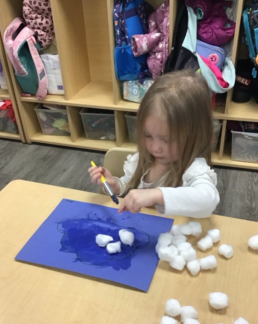 Young child paints cotton balls on blue paper at a table in a classroom.