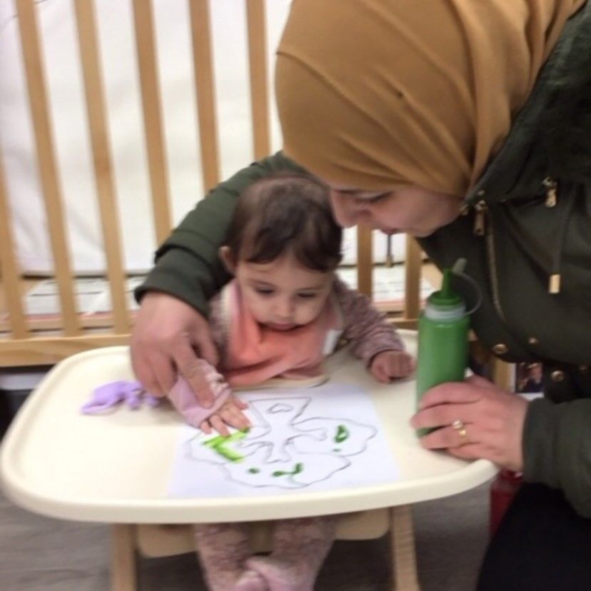 Woman helps a baby paint with green paint on a paper at a table.