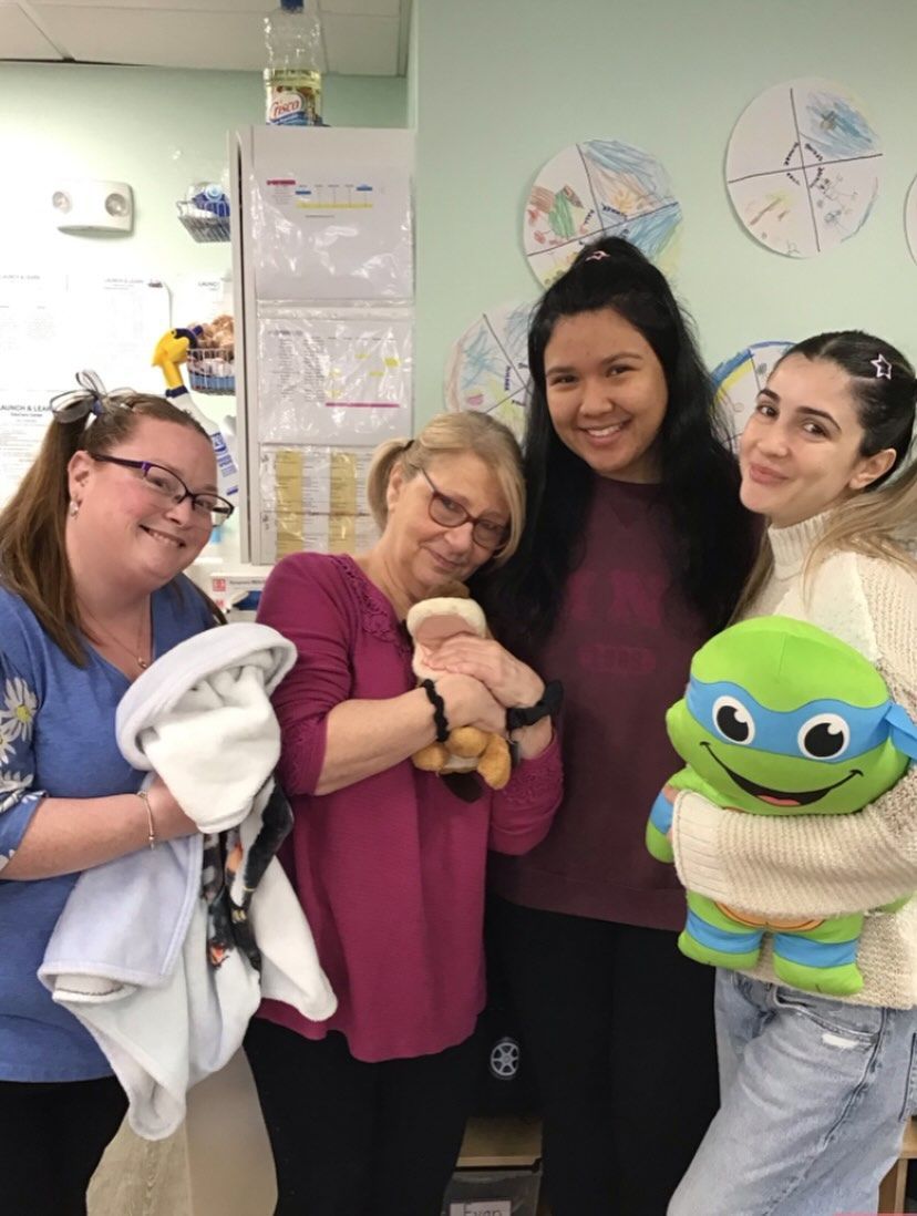 Four people holding baby items and stuffed toys, smiling in a room with pastel walls and decorations.