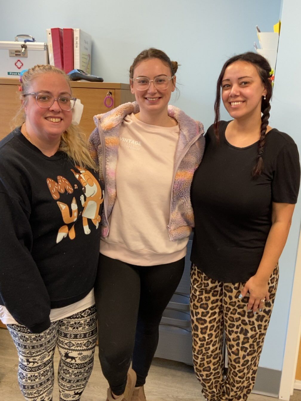 Three women smiling, standing close together indoors. One in black sweatshirt with fox graphic and patterned pants, one in a pink sweatshirt and black pants, and one in a black shirt and leopard-print pants.