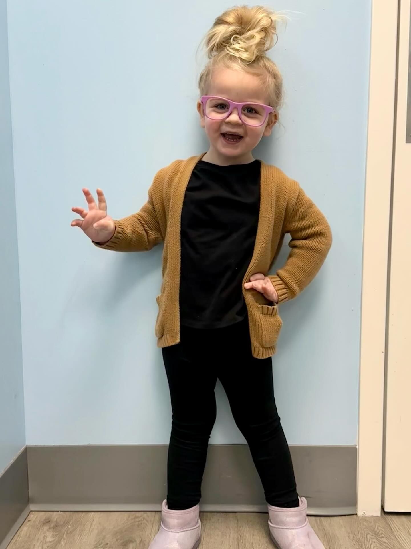 Blonde toddler with glasses and hair bun smiles, wearing black shirt, leggings, tan cardigan, and pink boots.