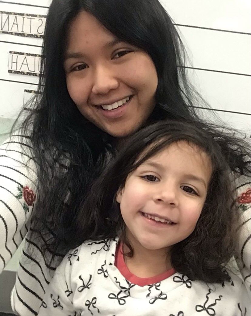 Woman and young child smiling, close-up. Both have dark hair, wearing patterned tops, possibly indoors.