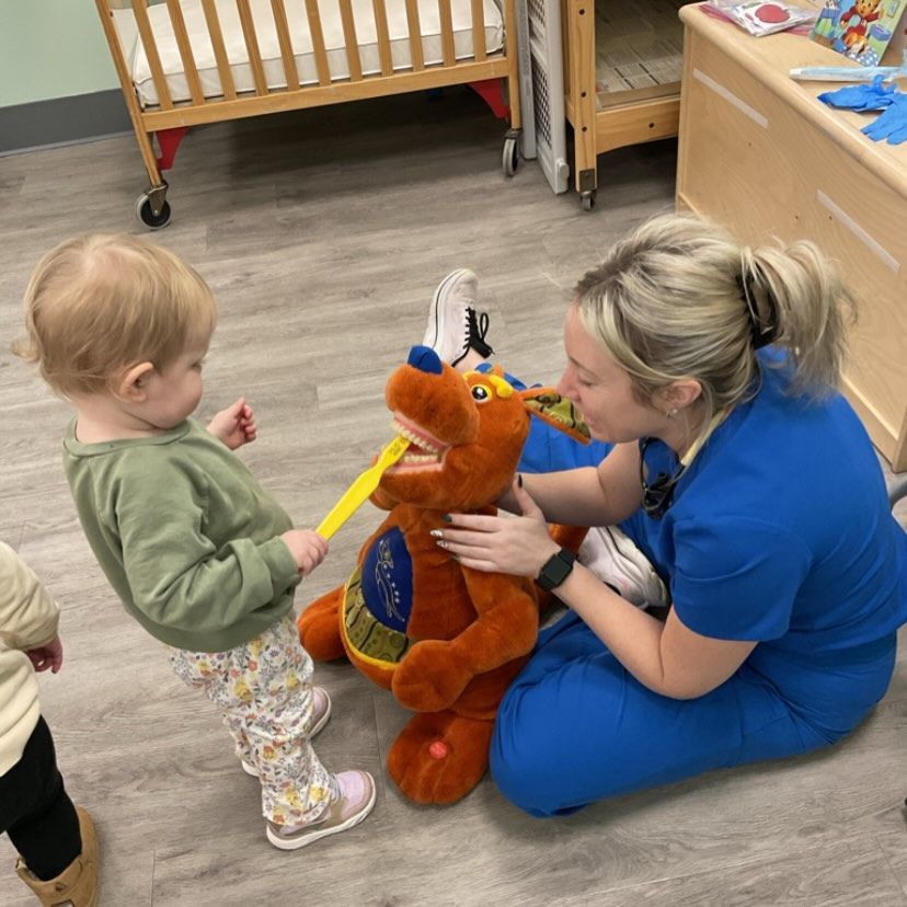 A child brushes the teeth of a plush toy with the help of a woman in blue scrubs.
