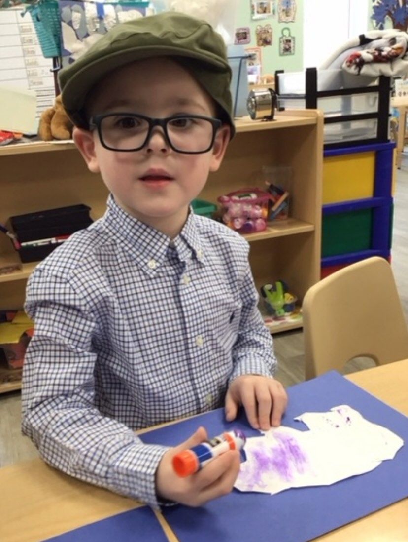 Boy in cap and glasses coloring a shape with a glue stick at a table in a classroom.