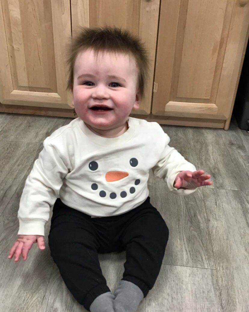 Smiling baby in snowman shirt and black pants sitting on the floor.