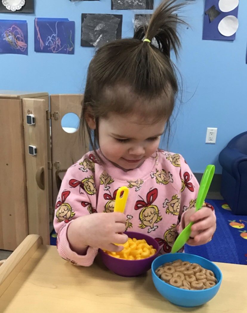 Child playing with toy food in bowls, smiling, and holding utensils. Blue wall background.