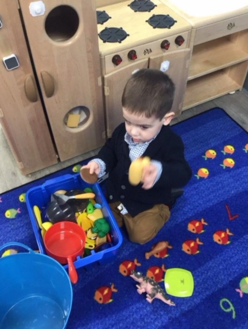 Child playing with toys in a play kitchen. Blue rug, toy fish, and colorful objects.