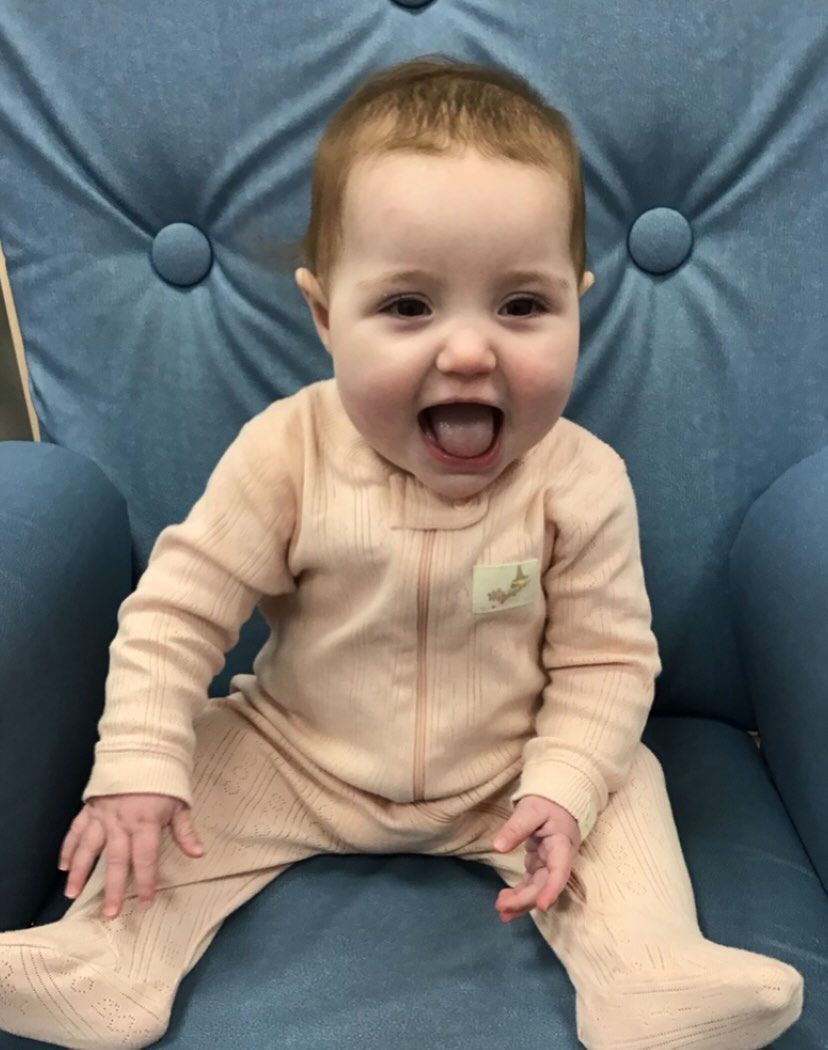Baby in pink onesie smiles with tongue out, sitting in a blue button-backed chair.
