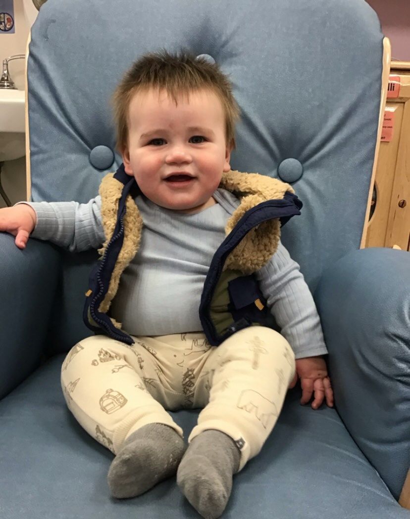 Baby sitting in blue armchair, wearing a vest, light blue shirt, patterned pants, and grey socks.