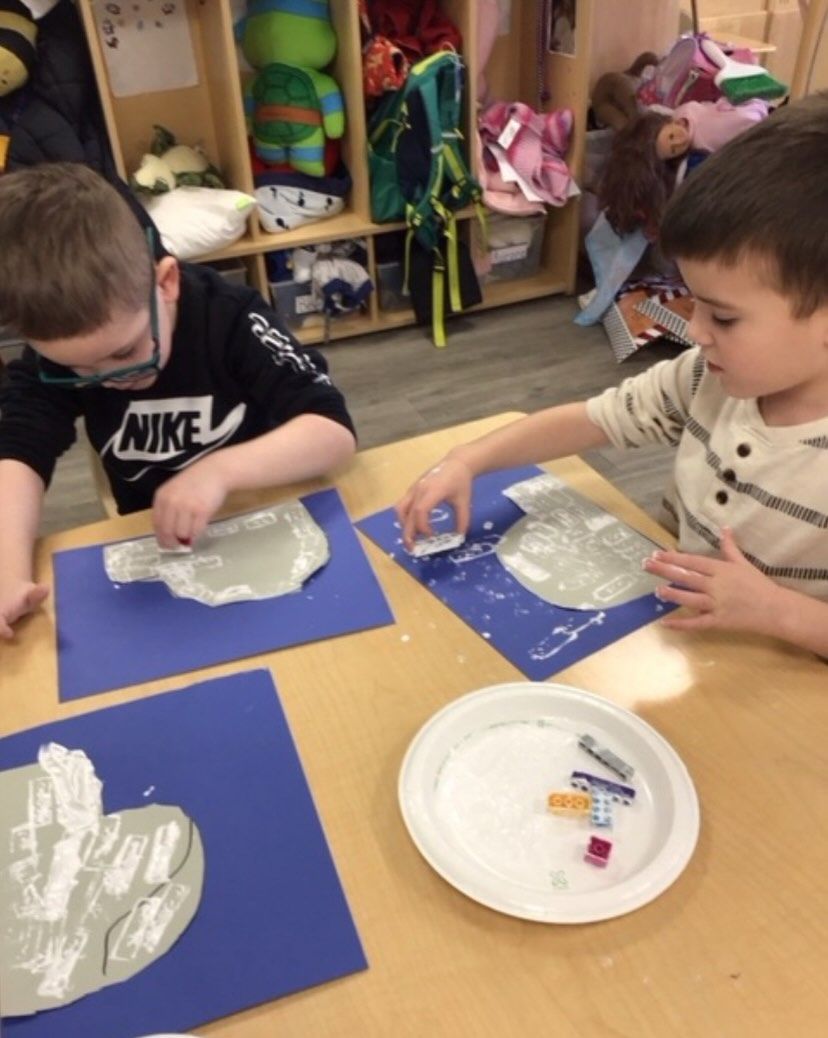 Two children painting gray shapes on blue paper at a table in a classroom.