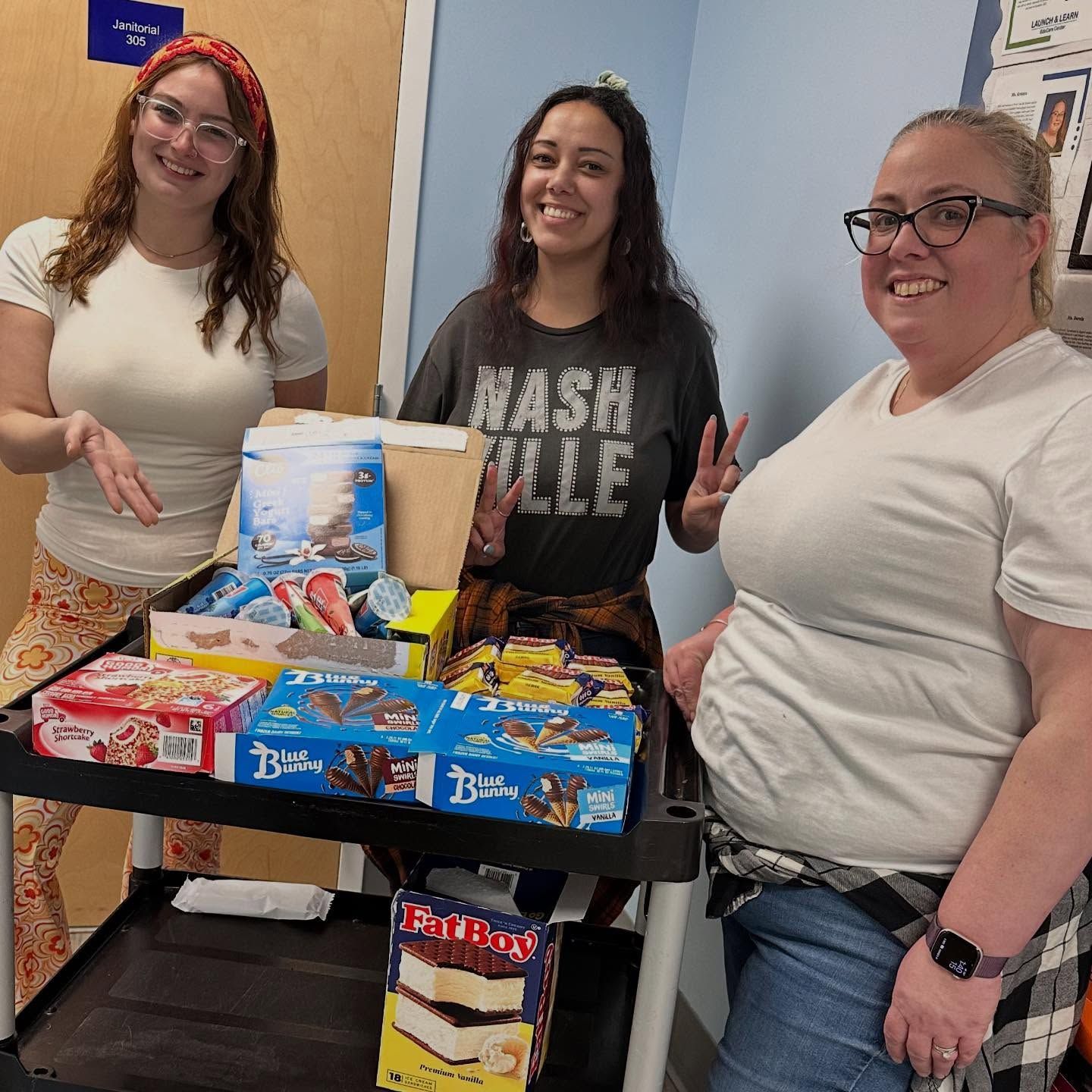 Three people stand near a cart of snacks. One points, one gives a peace sign, all smile.