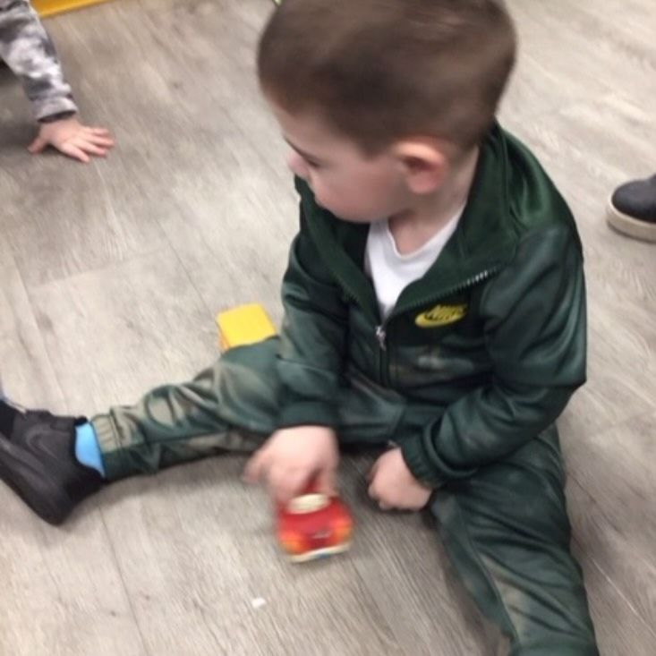 Young child in green tracksuit plays with a toy car on a wood floor.