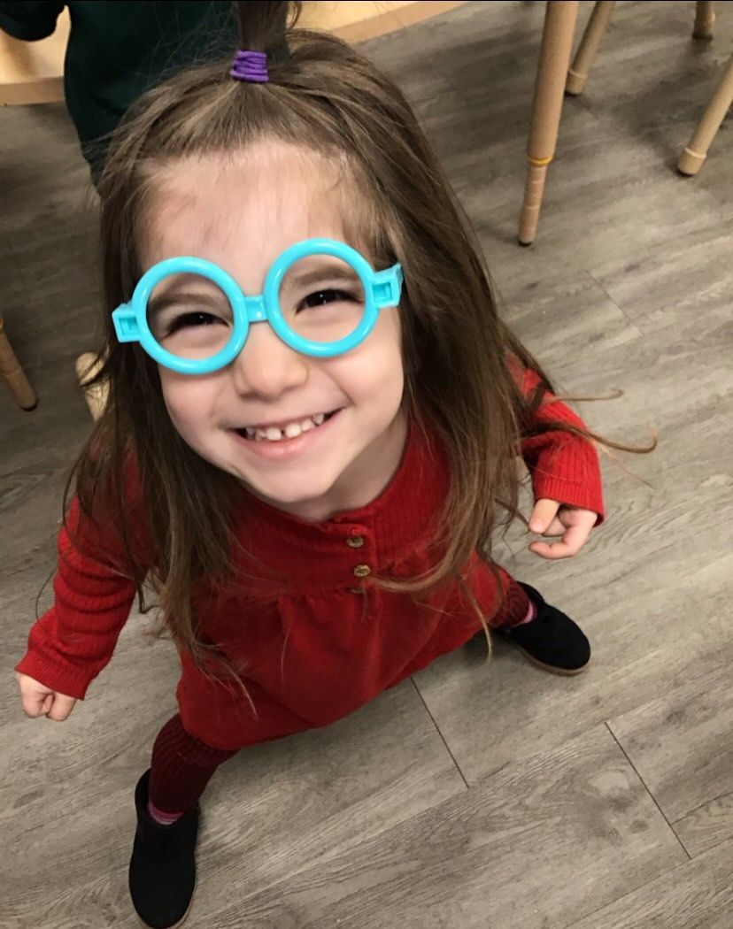Girl with long brown hair wearing light blue toy glasses and a red shirt smiles.