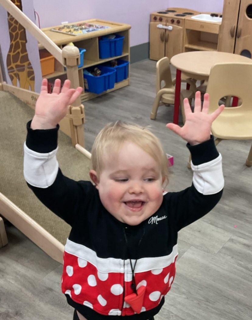 Child with arms raised, smiling, wearing a red polka-dot and black zip-up jacket. Indoors, with play furniture.
