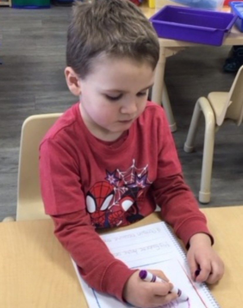 Boy in a red Spiderman shirt writing with a marker at a table.