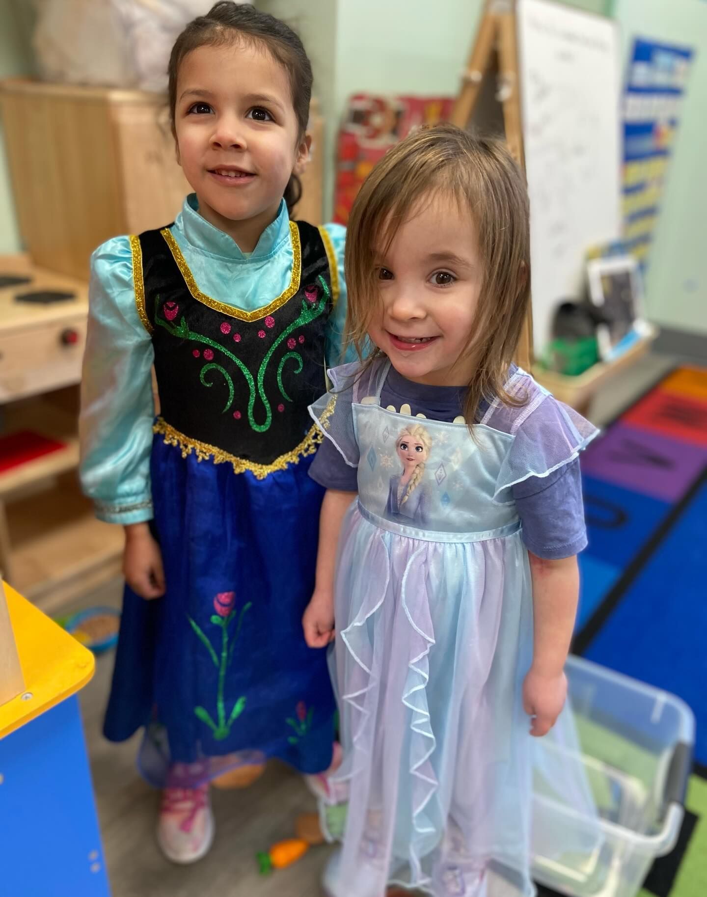 Two young children in princess costumes smile indoors. One wears blue, the other wears light blue.