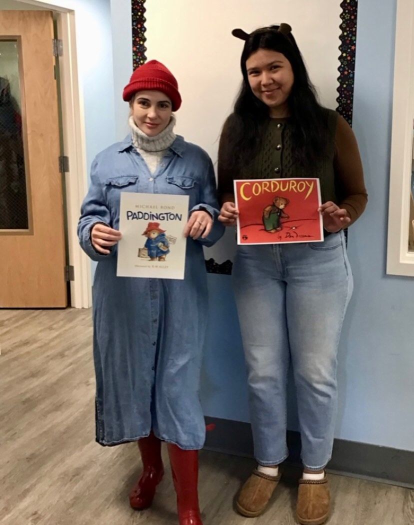 Two people dressed as characters from children's books, holding the book covers. One in a red hat and denim, the other in bear ears.