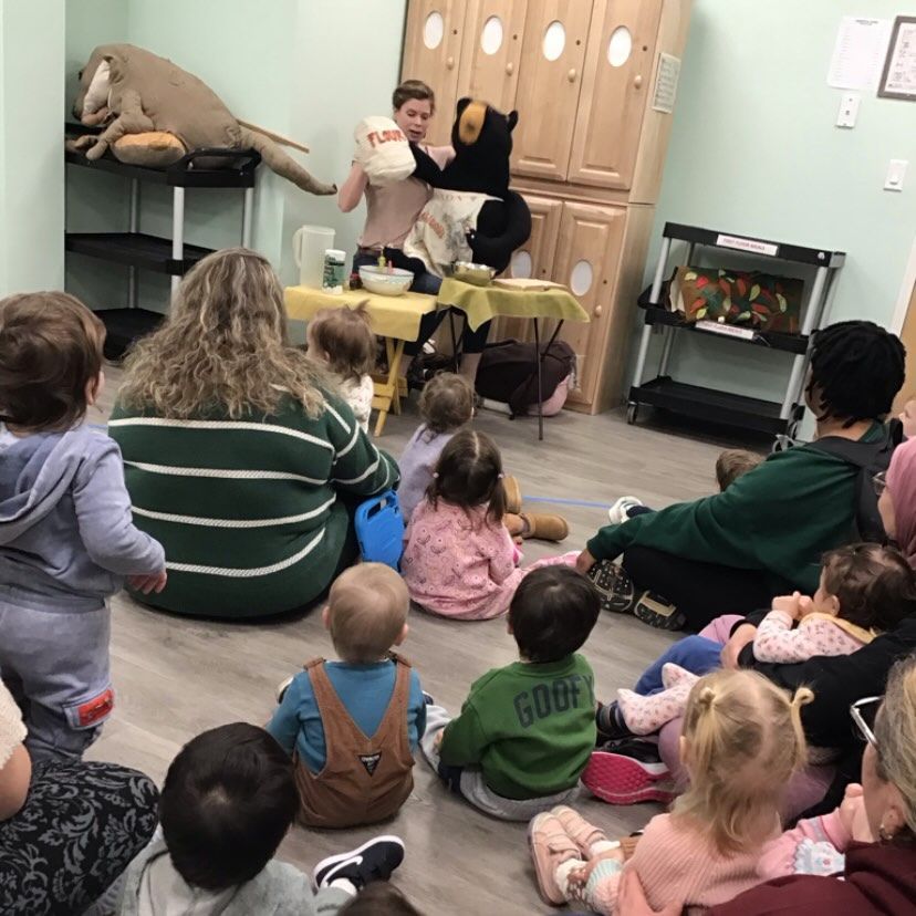 A person and bear puppet lead children in a cooking activity. Children sit on the floor, watching.